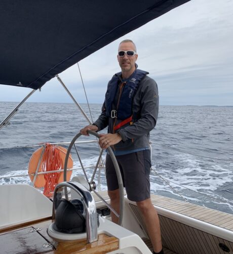 Skipper Christoph steuert eine Segelyacht über offenes Meer, trägt Sonnenbrille und Rettungsweste, unter einem dunklen Cockpitdach.“